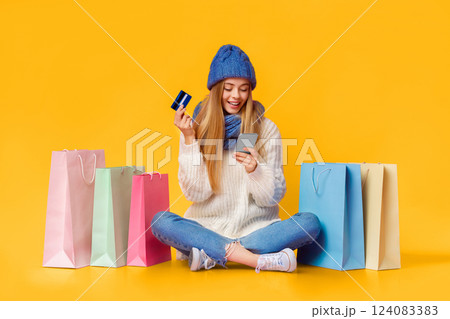 Pretty blonde girl in blue knitted hat using smartphone and credit card for online shopping, sitting on floor among shopping bags, pink background, copy space Pretty blonde girl in blue knitted hat using smartphone and credit card for online shopping, sitting on floor among shopping bags, pink background, copy space 124083383