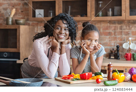 African sweet mom and daughter leaning on elbows and smiling, cooking healthy meal together at kitchen, free space 124083444