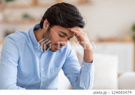 Arab man with dark hair is sitting in a chair indoors. He has a stressed expression on his face. He is holding his hand to his forehead, as if he is experiencing a headache or feeling overwhelmed. Arab man with dark hair is sitting in a chair indoors. He has a stressed expression on his face. He is holding his hand to his forehead, as if he is experiencing a headache or feeling overwhelmed. 124083474