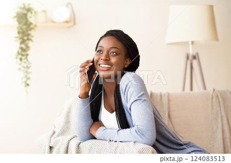 Young african woman talking on mobile phone with friend while sitting on comfortable couch at home, copy space 124083513