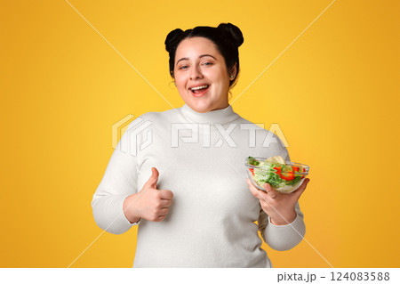 Healthy Eating. Happy Plus Size Girl Holding Bowl With Fresh Vegetable Salad And Showing Thumb Up Gesture, Standing Over Yellow Background, Free Space Healthy Eating. Happy Plus Size Girl Holding Bowl With Fresh Vegetable Salad And Showing Thumb Up Gesture, Standing Over Yellow Background, Free Space 124083588