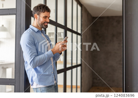 A man wearing a blue button-up shirt and jeans stands near a large glass door in a modern apartment. He is looking down at his smartphone and smiling, copy space 124083696