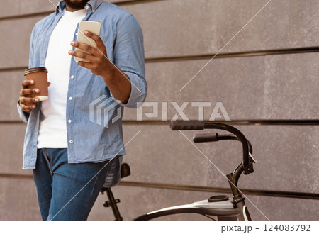 Unrecognizable black man with bicycle browsing internet on smartphone and drinking takeaway coffee outdoors 124083792
