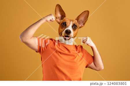A dog with a brown and white coat, wearing an orange t-shirt, stands against a yellow background with both arms flexed to show off its biceps. The dog's tongue is sticking out 124083801