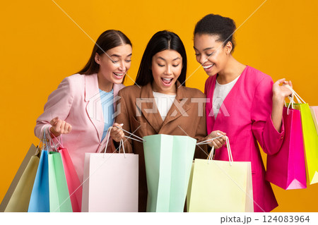 Shopping Concept. Three Excited Girls Looking Into Shopper Bags Standing On Yellow Background. Studio Shot Shopping Concept. Three Excited Girls Looking Into Shopper Bags Standing On Yellow Background. Studio Shot 124083964