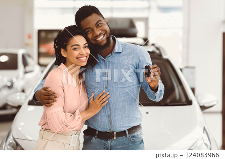 Car Buyers. African Spouses Showing New Vehicle Key To Camera Smiling And Hugging Near Auto In Dealership Showroom. Car Buyers. African Spouses Showing New Vehicle Key To Camera Smiling And Hugging Near Auto In Dealership Showroom. 124083966