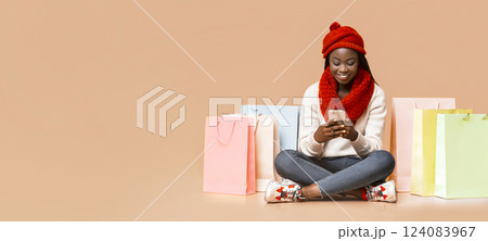 A cheerful young black woman wearing a red hat and scarf sits on the floor, happily using her smartphone. Colorful shopping bags are arranged around her, copy space A cheerful young black woman wearing a red hat and scarf sits on the floor, happily using her smartphone. Colorful shopping bags are arranged around her, copy space 124083967