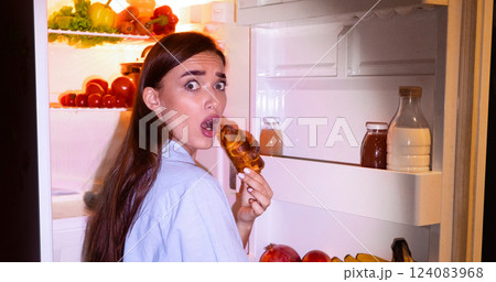 Failure of diet. Young woman eating junk croissant near opened fridge late in night, free space Failure of diet. Young woman eating junk croissant near opened fridge late in night, free space 124083968