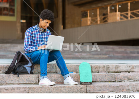 A teen black guy with short, curly hair sits on a set of concrete steps in front of a building, using a laptop computer. A black backpack sits beside him, and a teal skateboard is resting on the step A teen black guy with short, curly hair sits on a set of concrete steps in front of a building, using a laptop computer. A black backpack sits beside him, and a teal skateboard is resting on the step 124083970