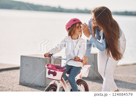 Woman is helping the little girl. Mother with her young daughter is with bicycle outdoors together Woman is helping the little girl. Mother with her young daughter is with bicycle outdoors together 124084579