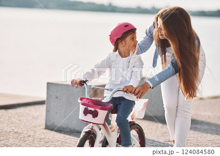 Woman is helping the little girl. Mother with her young daughter is with bicycle outdoors together Woman is helping the little girl. Mother with her young daughter is with bicycle outdoors together 124084580