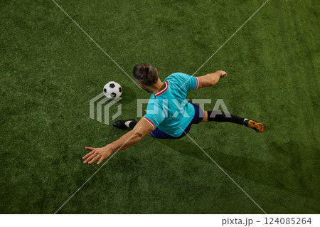 Top-down perspective of skilled football player executing powerful pass, leg fully extended, ball rolling on stadium grass. 124085264