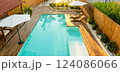 Modern backyard with turquoise swimming pool, wooden deck, umbrellas, and lounge chairs in a serene setting 124086066