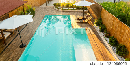 Modern backyard with turquoise swimming pool, wooden deck, umbrellas, and lounge chairs in a serene setting 124086066