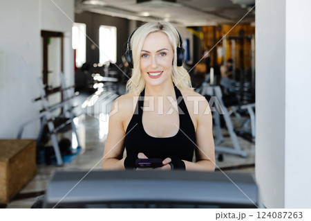 Athletic woman using smartphone and wearing headphones on treadmill in gym 124087263