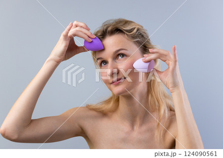 A Young Woman Engaged in Makeup Application Using Beauty Blenders for Flawless Skin While Gazing Thoughtfully in a Light Gray Background Setting 124090561