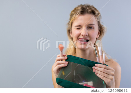 Playful Woman Winking and Sticking Out Her Tongue Holding a Makeup Bag with Brushes Against a Light Blue Background in a Fun and Cheerful Atmosphere 124090651