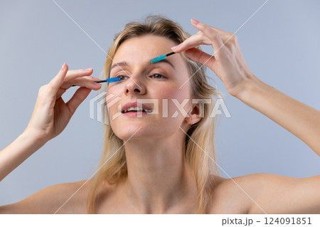 Woman Applying Makeup with Two Different Brushes on Her Eyelashes While Smiling Brightly at the Camera in a Soft Gray Background Capturing a Carefree Beauty Moment 124091851