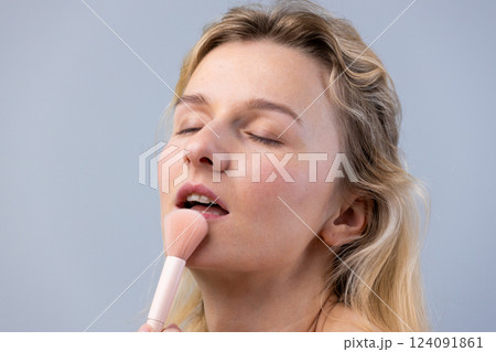 Captivating Beauty: Young Woman Engages in a Delicate Makeup Application with a Soft Brush Against a Minimalist Background, Highlighting Elegance and Radiance 124091861