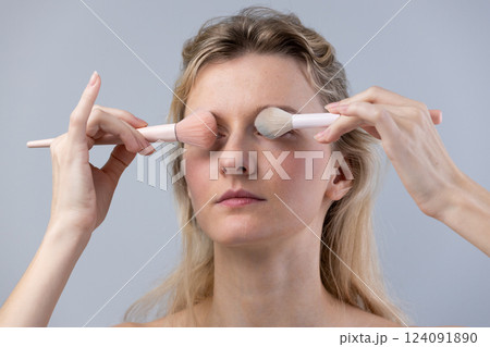 A Young Woman Applying Makeup with Elegant Brushes on a Light Gray Background: The Art of Skincare and Cosmetic Enhancement A Young Woman Applying Makeup with Elegant Brushes on a Light Gray Background: The Art of Skincare and Cosmetic Enhancement 124091890