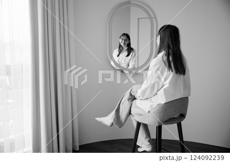 Beautiful girl looking at herself in mirror and smiling. Pretty female checking herself. Cute girl sitting in chair opposite to mirror at home. Black and white image. Beautiful girl looking at herself in mirror and smiling. Pretty female checking herself. Cute girl sitting in chair opposite to mirror at home. Black and white image. 124092239