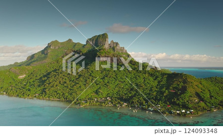 Majestic green mountain rising above Bora Bora turquoise waters, lush landscape under bright blue sky. French Polynesia tropical paradise. Exotic travel vacation getaway. Aerial drone flight zoom in 124093348