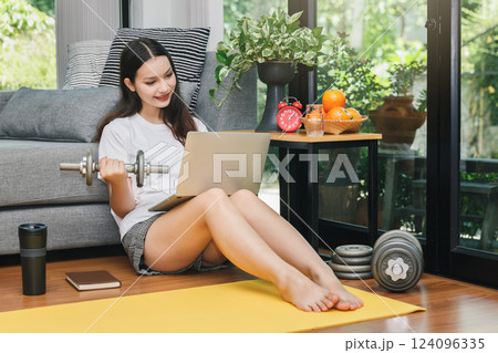 Beautiful young woman with dumbbell watching fitness video tutorials online on laptop while sit down on floor in living room. 124096335