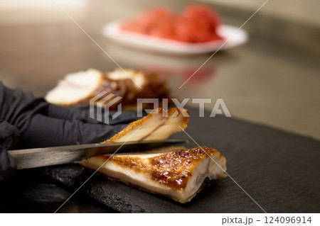 A chef with black gloves skillfully slices tender roasted meat on a slate board. Fresh ingredients can be seen in the background, enhancing the culinary atmosphere A chef with black gloves skillfully slices tender roasted meat on a slate board. Fresh ingredients can be seen in the background, enhancing the culinary atmosphere 124096914