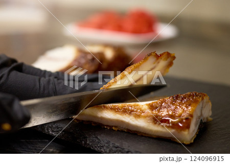 A skilled chef slices through crispy pork in a modern kitchen. The juicy meat is complemented by vibrant garnishes placed on a white plate, showcasing culinary precision and artistry A skilled chef slices through crispy pork in a modern kitchen. The juicy meat is complemented by vibrant garnishes placed on a white plate, showcasing culinary precision and artistry 124096915