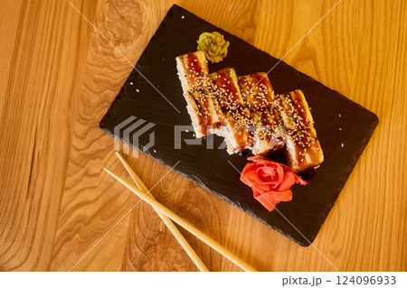 Grilled eel sushi arranged neatly on a black slate board with sesame seeds, garnished with wasabi, ginger, and decorative rose on wooden table 124096933