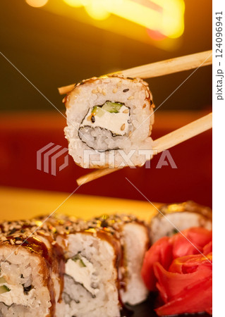 A close-up of a sushi roll being picked up with chopsticks in a lively restaurant. The colorful sushi features rice, vegetables, and a touch of sauce, set against a softly lit background 124096941