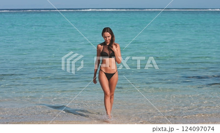 Young woman enjoying a sunny, tropical vacation walks out of the ocean onto a pristine beach, exuding happiness and relaxation 124097074