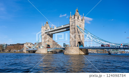 Tower Bridge stands majestically against a clear blue sky, showcasing its iconic architecture. People can be seen enjoying the waterfront, while boats navigate the Thames River. 124098784