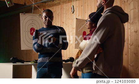 Shooting gallery safety officer demonstrating how to safely reload pistol and cock gun to clients, supervising range activities. Firing range expert teaching proper weapon handling and usage, camera A 124103080