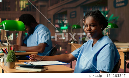African american med school pupil studies pharmacology notes in a library, preparing for exams and advancing their healthcare career. Girl in scrubs works hard on medical school success. Camera B. 124103245
