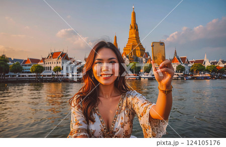 Woman is smiling and taking a picture of herself with a cell phone. She is wearing a white and yellow dress and is standing in front of a large building. The water in the background is calm and 124105176