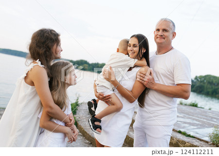 Family of five is standing on pier by the water, with woman holding baby. Atmosphere is warm and friendly 124112211