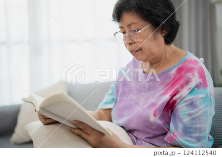 Senior Asian Woman Enjoying a Peaceful Reading Session on a Cozy Sofa in a Bright Living Room with Natural Light and Minimalist Decor Senior Asian Woman Enjoying a Peaceful Reading Session on a Cozy Sofa in a Bright Living Room with Natural Light and Minimalist Decor 124112540