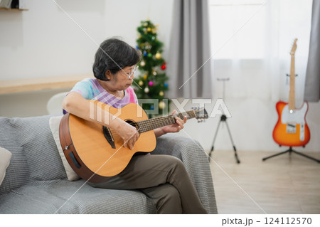 Senior Asian woman playing acoustic guitar at home, enjoying music, dressed in colorful shirt, sitting on couch with natural light and cozy interior Senior Asian woman playing acoustic guitar at home, enjoying music, dressed in colorful shirt, sitting on couch with natural light and cozy interior 124112570