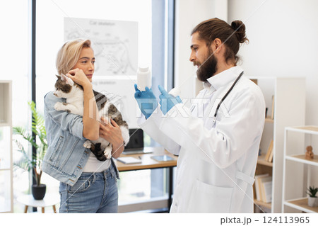 Vet explaining medication instructions to blonde young woman holding domestic tabby cat. Interaction depicting dedication to pet health in bright animal clinic setting. 124113965