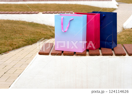 Three vibrant paper shopping bags standing on outdoor bench. Three vibrant paper shopping bags standing on outdoor bench. 124114626