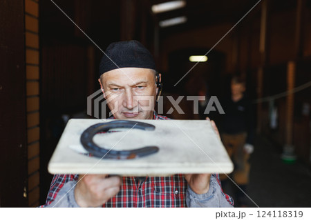 European busy blacksmith inspects horseshoe made for horse in stable European busy blacksmith inspects horseshoe made for horse in stable 124118319