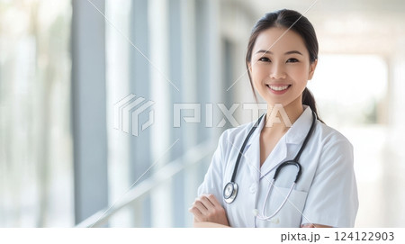 Confident Asian female doctor in a medical uniform, standing in a sunlit hospital corridor. She is smiling warmly, with soft natural lighting. The left side of the image is open for text placement Confident Asian female doctor in a medical uniform, standing in a sunlit hospital corridor. She is smiling warmly, with soft natural lighting. The left side of the image is open for text placement 124122903