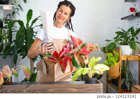 Woman unpacks a box with a parcel of potted flowers of red aglaonem - order wrapped tropical plants, delivery. Sale, safe shipment of plants from the store, packaging. Flower shop, home business Woman unpacks a box with a parcel of potted flowers of red aglaonem - order wrapped tropical plants, delivery. Sale, safe shipment of plants from the store, packaging. Flower shop, home business 124123241