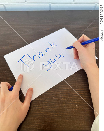 A close-up view of a white sheet of paper with the handwritten words Thank You placed on a dark wooden table, with two hands are nearby, one holding a blue marker 124123298