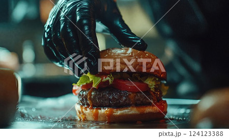 juicy burger being carefully assembled by a gloved hand. The burger has a toasted bun, fresh lettuce, tomato, and a thick patty with sauce dripping down, highlighting its appetizing appeal 124123388