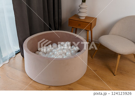 Modern Home Interior with Beige Soft Play Ball Pit Filled with White Plastic Balls Beside Minimalistic Wooden Side Table and Gray Chair Modern Home Interior with Beige Soft Play Ball Pit Filled with White Plastic Balls Beside Minimalistic Wooden Side Table and Gray Chair 124123926