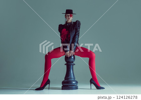 Fierce figure in structured black clothing and dramatic red details straddles oversized chess piece. Fierce figure in structured black clothing and dramatic red details straddles oversized chess piece. 124126278