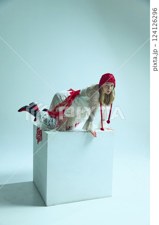 Young woman in white lace dress with red accents crawling on white cube, gazing intensely. Concept of surreal fashion Young woman in white lace dress with red accents crawling on white cube, gazing intensely. Concept of surreal fashion 124126296