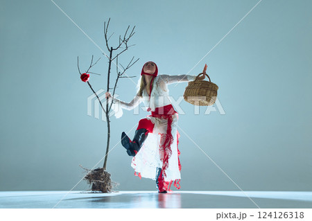 Woman in fluid motion, holding basket, posed beside bare tree with blooming rose, symbolizes fragile resilience and change. 124126318